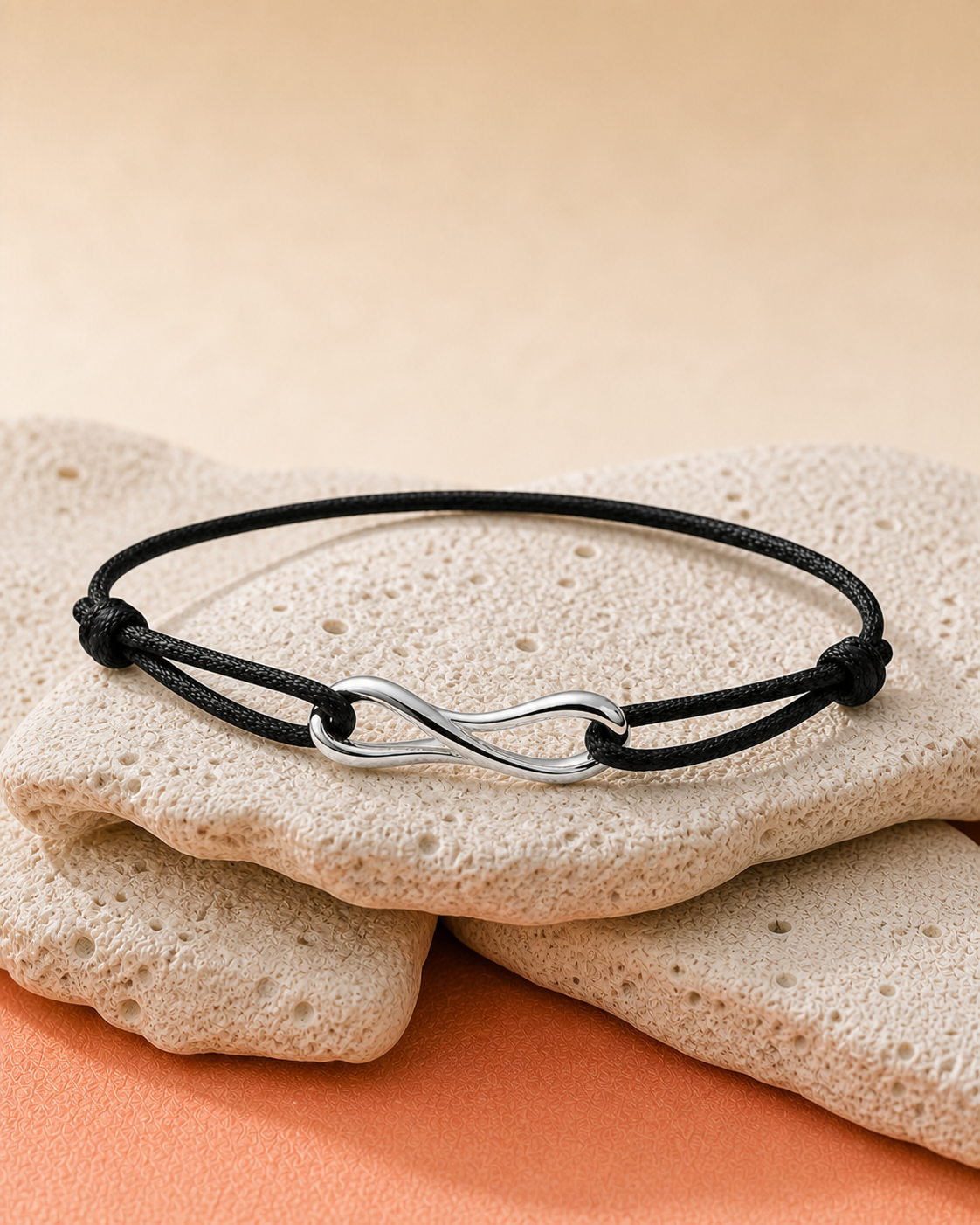 Coastal Rope Bracelet with black cord and a mirror-polished sterling-silver wave connector resting on pale coastal stone above a warm coral surface.